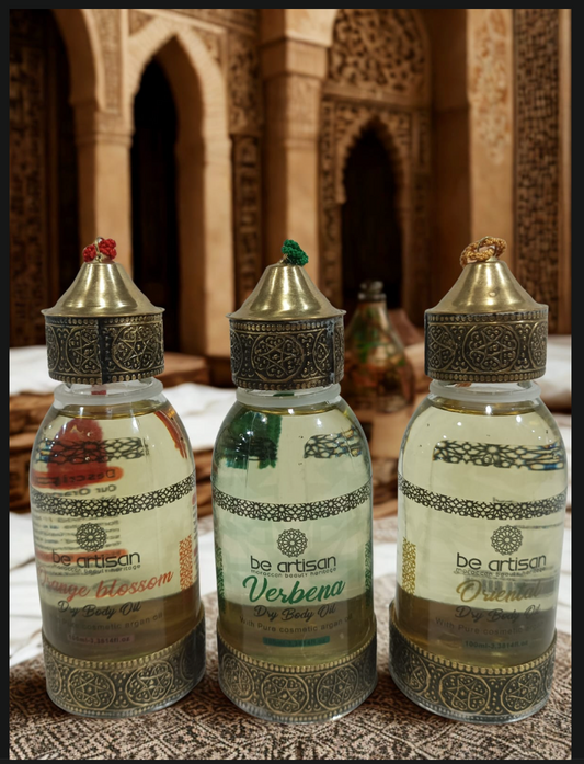 “Be Artisan Orange Blossom Dry Body Oil in ornate golden bottles with floral Moroccan detailing, front and back view on warm patterned fabric.”