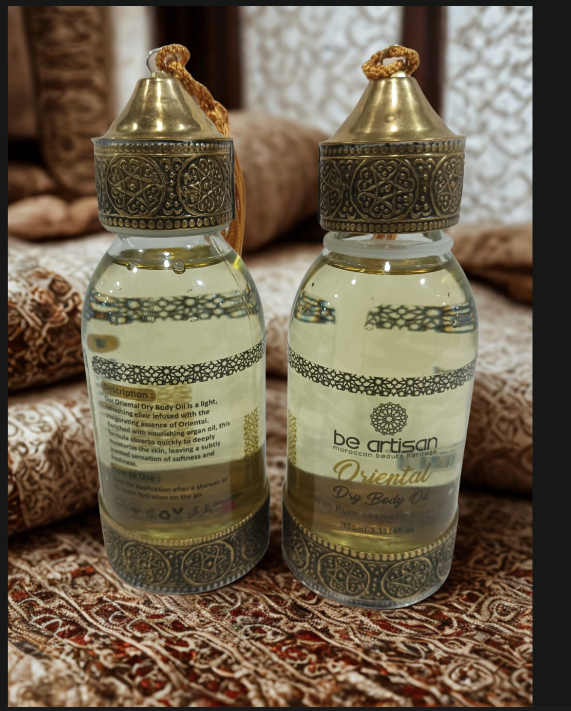“Be Artisan Oriental Dry Body Oil in ornate gold-topped bottles, front and back view, Moroccan-style design on patterned fabric background.”