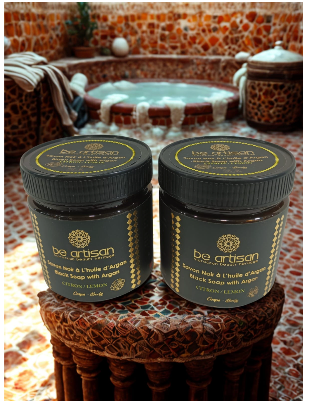 “Be Artisan Lemon & Argan Black Soap jars on Moroccan hammam tiles, traditional black soap with argan and lemon extract.”