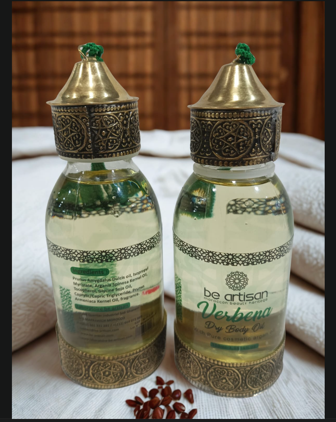 “Be Artisan Verbena Dry Body Oil in ornate gold-topped bottles, front and back view, Moroccan-style design on soft white towel background with seeds.”