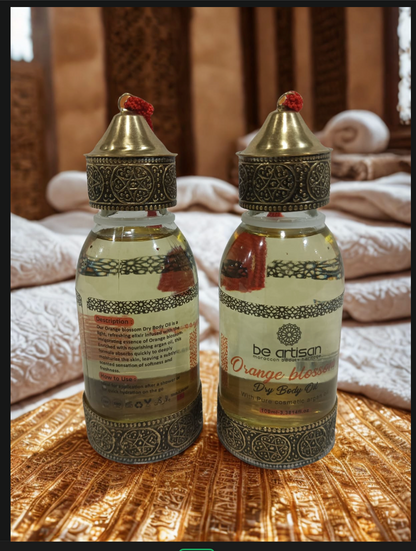 “Be Artisan Orange Blossom Dry Body Oil in ornate golden bottles with floral Moroccan detailing, front and back view on warm patterned fabric.”